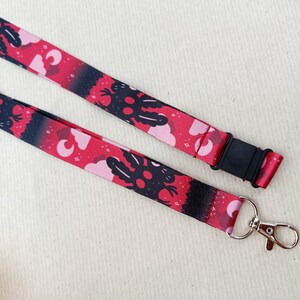May include: A red and black lanyard with a repeating pattern of a black creature with pink clouds and moons. The lanyard has a black plastic clip and a silver swivel hook.