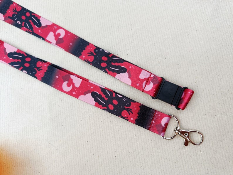 May include: A red and black lanyard with a repeating pattern of a black creature with pink clouds and moons. The lanyard has a black plastic clip and a silver swivel hook.
