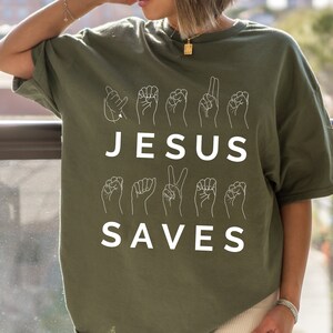 May include: A green t-shirt with the words "JESUS SAVES" in white lettering. The words are spelled out using American Sign Language hand gestures.
