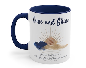 May include: White ceramic coffee mug with a navy blue rim and handle. The mug features a gold and blue watercolor sunrise with the text "Arise and Shine" and the scripture "for your light has come and the glory of the Lord has risen upon you" with the verse reference "ISAIAH 60:1".