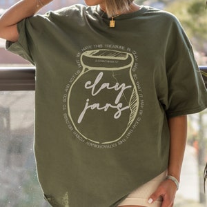 May include: Green t-shirt with a white graphic of a clay jar and the text "clay jars" and a Bible verse from 2 Corinthians 4:7. The text around the jar reads "We have this treasure in clay jars so that it may be clear that this extraordinary power belongs to God and not us."
