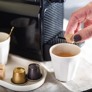 Peut inclure: Une tasse en céramique blanche avec un morceau de sucre brun ajouté. La tasse est posée sur une surface blanche avec une machine à café en arrière-plan. Il y a aussi deux dosettes de café sur la surface.