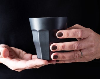 May include: A person's hand holding a black ceramic cup with a textured surface.