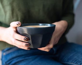 May include: A dark charcoal gray ceramic bowl with a faceted, geometric exterior design. The bowl has a wide, open top and a smooth matte finish, featuring a distinct angular shape that tapers toward the base, held firmly in place.