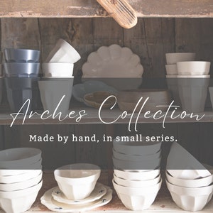 May include: A wooden shelf displays a collection of ceramic tableware. The "Arches Collection" includes white, grey, and navy blue cups, bowls, and plates. The text "Made by hand, in small series" is also visible.