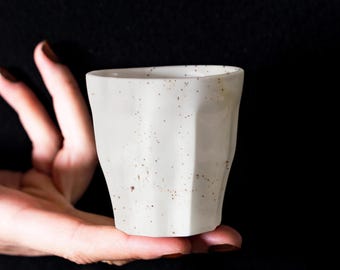 May include: A white ceramic cup with a speckled brown glaze. The cup has a unique, faceted design.