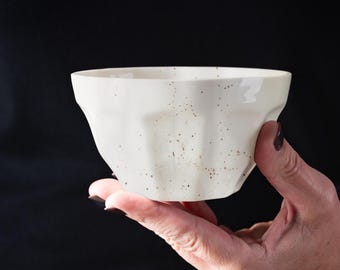 May include: A white ceramic bowl with a speckled glaze. The bowl has a fluted design and is held in a hand.