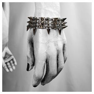 K&ouml;nnte beinhalten: Ein silberfarbenes Armband mit mehreren Reihen von Metallspitzen. Das Armband wird an einer wei&szlig;en Hand mit schwarzem Nagellack getragen. Das Bild ist in Schwarzwei&szlig;, mit einer zweiten Hand im Hintergrund sichtbar.
