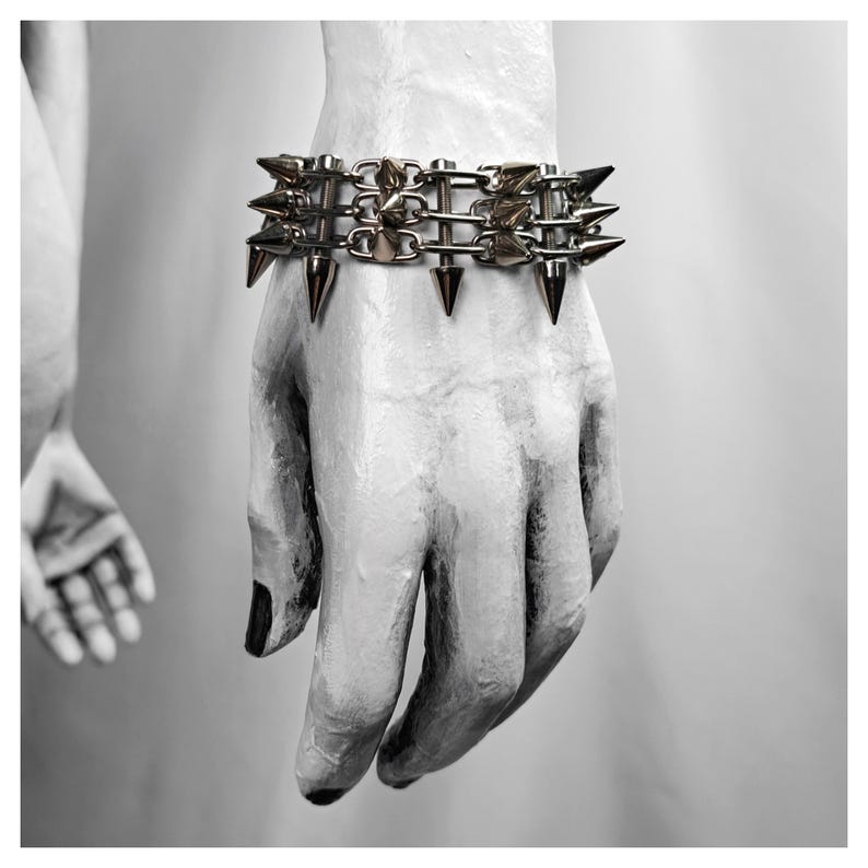 K&ouml;nnte beinhalten: Ein silberfarbenes Armband mit mehreren Reihen von Metallspitzen. Das Armband wird an einer wei&szlig;en Hand mit schwarzem Nagellack getragen. Das Bild ist in Schwarzwei&szlig;, mit einer zweiten Hand im Hintergrund sichtbar.