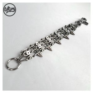 K&ouml;nnte beinhalten: Ein silberfarbenes Kettenarmband mit drei Reihen von Kettengliedern und spitzen Stacheln. Das Armband hat einen gro&szlig;en Ring an einem Ende und einen Verschluss am anderen. Der Hintergrund ist eine schlichte wei&szlig;e Oberfl&auml;che.