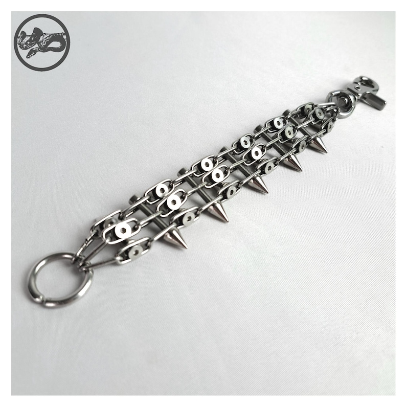 K&ouml;nnte beinhalten: Ein silberfarbenes Kettenarmband mit drei Reihen von Kettengliedern und spitzen Stacheln. Das Armband hat einen gro&szlig;en Ring an einem Ende und einen Verschluss am anderen. Der Hintergrund ist eine schlichte wei&szlig;e Oberfl&auml;che.