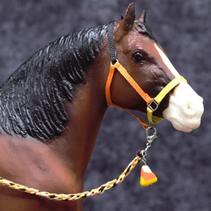 May include: A brown and white horse figurine with a black mane and a white stripe on its face. It wears a yellow and orange halter and a matching lead rope with a candy corn charm. The horse is a decorative item.