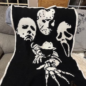 May include: Black and white crocheted blanket featuring iconic horror movie characters: Michael Myers, Jason Voorhees, Ghostface, and Freddy Krueger. The blanket has a white border and is displayed on a gray couch.