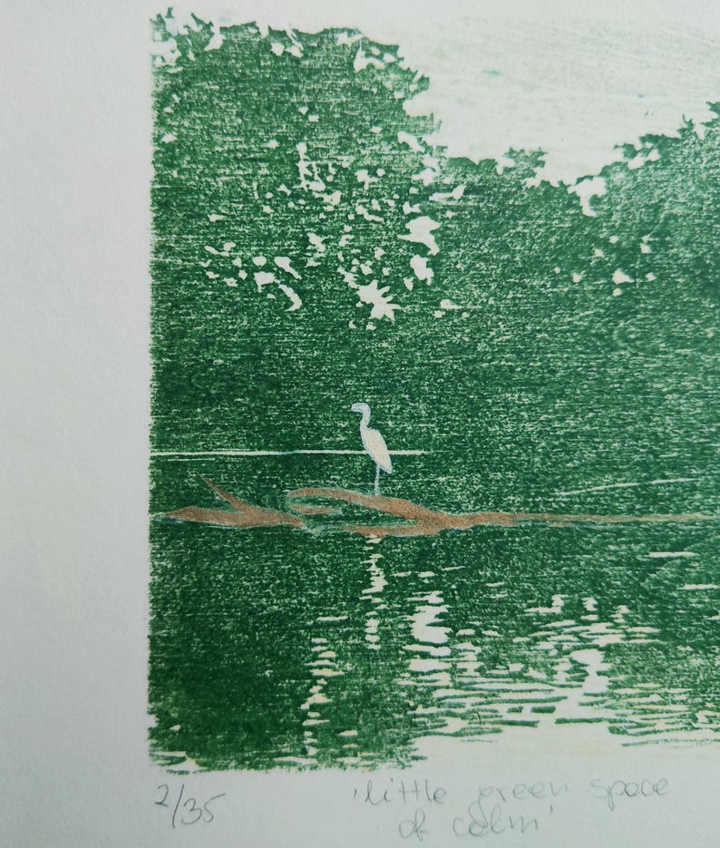 May include: A green-toned print on paper featuring a white heron standing on a brown log in water, with trees in the background. The print is numbered 2/35 and titled 'little green space of calm' in handwritten text at the bottom edge.