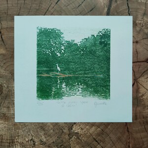 May include: A square green ink print on white paper depicting a white heron standing on a log in a lake surrounded by trees. The print features the handwritten text 2/35, little meer space of calm, and a signature in the bottom margin area.