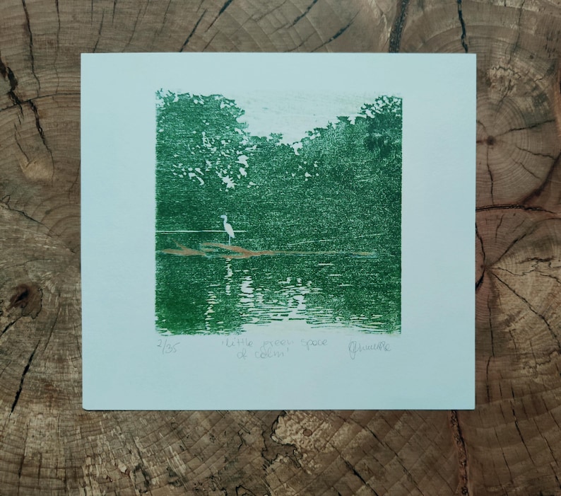 May include: A square green ink print on white paper depicting a white heron standing on a log in a lake surrounded by trees. The print features the handwritten text 2/35, little meer space of calm, and a signature in the bottom margin area.