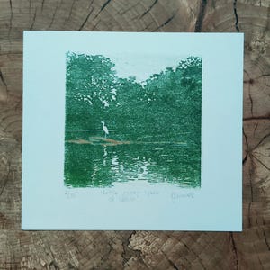 May include: A square art print on white paper featuring a green ink block print of a white heron standing on a log in a lake surrounded by trees. The print includes the handwritten text 2/35, little green space of calm, and a signature.