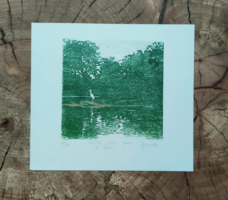 May include: A square art print on white paper featuring a green ink block print of a white heron standing on a log in a lake surrounded by trees. The print includes the handwritten text 2/35, little green space of calm, and a signature.