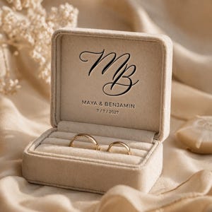 Puede incluir: Una caja de anillos de terciopelo beige abierta, que revela dos alianzas doradas. La caja lleva las iniciales "MB" y los nombres "MAYA & BENJAMIN" con la fecha "7/7/2027". El fondo es una tela suave de color crema.