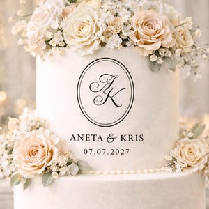 Può includere: Una torta nuziale bianca a due piani decorata con rose chiare e verde. La torta presenta le iniziali "AK" all'interno di una cornice circolare nera, con i nomi "ANETA & KRIS" e la data "07.07.2027".