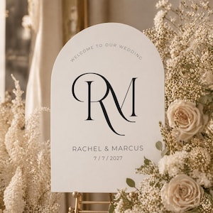May include: A cream-colored wedding welcome sign with an arched top. The sign features the initials "RM" in elegant black script, along with the names "Rachel & Marcus" and the date "7/7/2027". The sign is surrounded by floral arrangements.
