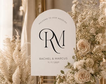 May include: A cream-colored wedding welcome sign with an arched top. The sign features the initials "RM" in elegant black script, along with the names "Rachel & Marcus" and the date "7/7/2027". The sign is surrounded by floral arrangements.