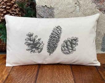 May include: A white rectangular pillow with a black and white illustration of three pine cones. The pine cones are arranged horizontally across the pillow.