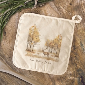 May include: Cream-colored pot holder with a scenic watercolor print of a deer in a forest with yellow and brown trees. The text "Sun Valley, Idaho" is printed at the bottom. The pot holder has a loop for hanging.