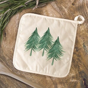 May include: Off-white square pot holder with a loop for hanging. The pot holder features a watercolor design of three green pine trees. The background is a wooden surface with a sprig of rosemary.