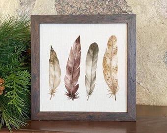 May include: A framed artwork featuring watercolor illustrations of five feathers in shades of brown, beige, and tan. The feathers are arranged vertically against a white background, enclosed in a dark brown wooden frame. The artwork evokes a rustic, natural theme.