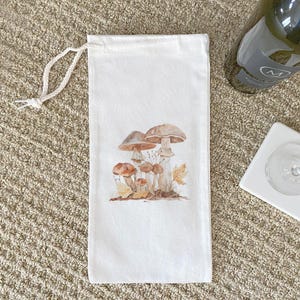 May include: A white canvas wine bottle bag featuring a watercolor illustration of several brown and tan mushrooms with autumn leaves. The bag has a drawstring closure and is designed to hold a standard wine bottle.