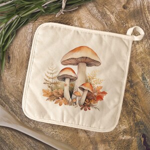 May include: Cream-colored square pot holder featuring a watercolor illustration of several mushrooms with orange caps and brown stems. The mushrooms are surrounded by autumn leaves and ferns. A loop is attached for hanging.
