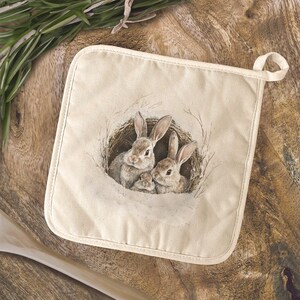 May include: Off-white square pot holder featuring a watercolor illustration of three rabbits nestled in a nest. The rabbits are shades of brown and gray. The pot holder has a loop for hanging.