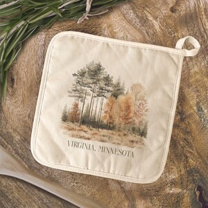 May include: Off-white pot holder with a watercolor illustration of a forest scene in shades of green, brown, and orange. The text "VIRGINIA, MINNESOTA" is printed at the bottom. The pot holder has a loop for hanging.