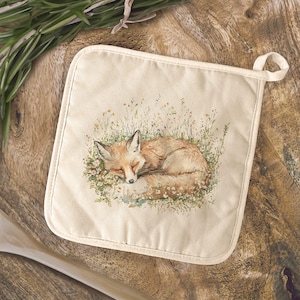 May include: A square, off-white pot holder featuring a watercolor illustration of a sleeping fox curled up in a bed of wildflowers. The fox has orange fur and closed eyes. A loop is attached for hanging.