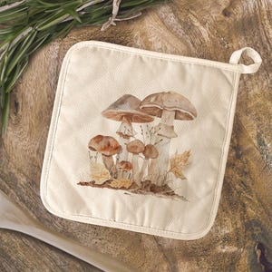 May include: Off-white square pot holder featuring a watercolor illustration of various brown mushrooms and autumn leaves. The pot holder has a loop for hanging and is made of a thick, durable fabric. The design evokes a natural, earthy aesthetic.