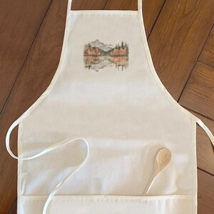May include: A white apron with a landscape design featuring mountains, trees, and a lake. A wooden spoon rests in the front pocket. The apron has a neck loop and waist ties.