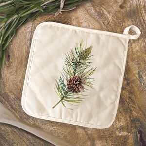 May include: A cream-colored cotton pot holder with a watercolor illustration of a pine branch with a brown pine cone.