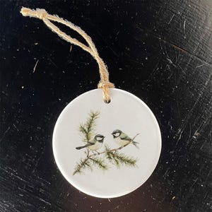 May include: Round white ceramic ornament featuring two small birds perched on a branch with green needles. The birds have gray and white feathers. A natural twine loop is attached for hanging.