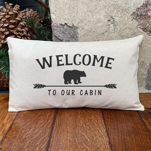 May include: Rectangular cream-colored pillow with the words "WELCOME TO OUR CABIN" in black. A black bear silhouette is centered above an arrow design. The pillow is on a wooden surface.