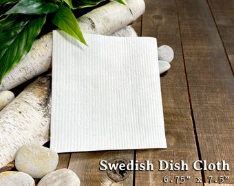 May include: A white Swedish dish cloth, measuring 6.75" x 7.5", is displayed on a wooden surface. The cloth is made of 70% cellulose and 30% cotton, and is reusable and machine washable. The text "Swedish Dish Cloth" is visible.