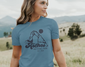 May include: A blue t-shirt with a graphic of Delicate Arch in Arches National Park, Utah. The text "Arches National Park Est. 1929" is printed below the arch.