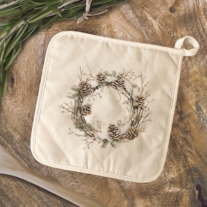 May include: Off-white square pot holder with a loop for hanging. The center features a detailed illustration of a wreath made of pinecones, twigs, and greenery. The pot holder is on a wooden surface.