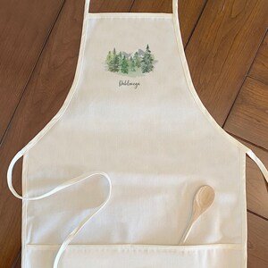 May include: White cotton apron with a green watercolor mountain scene and the word "Dabblemore" printed on it. The apron has a large pocket and a wooden spoon is in the pocket.