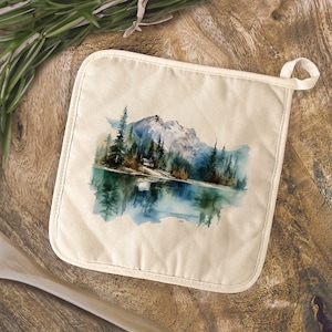 May include: A white cotton pot holder with a watercolor painting of a mountain lake scene. The painting features a small cabin nestled among the trees on the shore of the lake, with a mountain range in the background.
