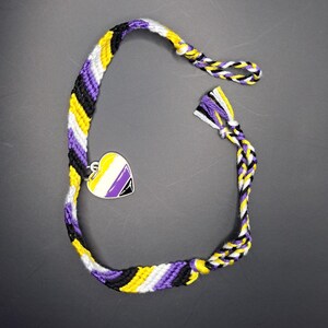 May include: A woven friendship bracelet featuring diagonal stripes in yellow, white, purple, and black thread. The bracelet includes a heart-shaped metal charm with an enamel inlay matching the yellow, white, purple, and black color scheme.