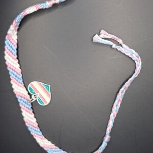 May include: A handmade friendship bracelet featuring a diagonal striped pattern in light blue, white, and light pink embroidery thread. A silver-toned metal heart-shaped charm with enamel stripes in matching light blue, white, and pink is attached.