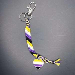 May include: A purple, yellow, and black friendship bracelet with a heart-shaped charm. The charm is also purple, yellow, and black.