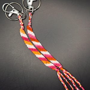 Op de afbeelding: Twee zilveren sleutelhangers met een roze, oranje en witte vriendschapsband.