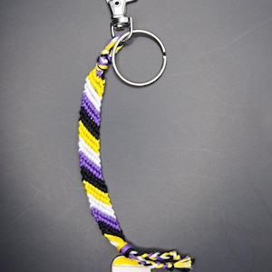 May include: A keychain with a purple, yellow, white, and black woven strap and a heart-shaped charm with the same colors. The charm is attached to a silver key ring.